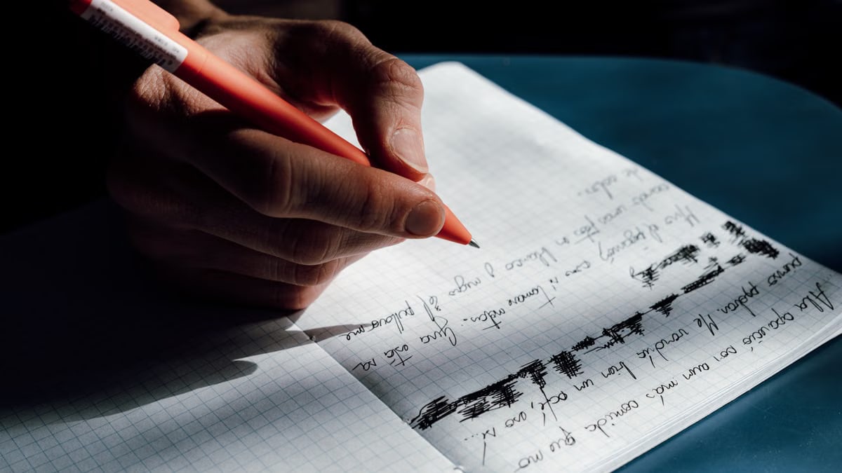 A hand writing with a red pen on a grid notebook, capturing thoughtful detail.